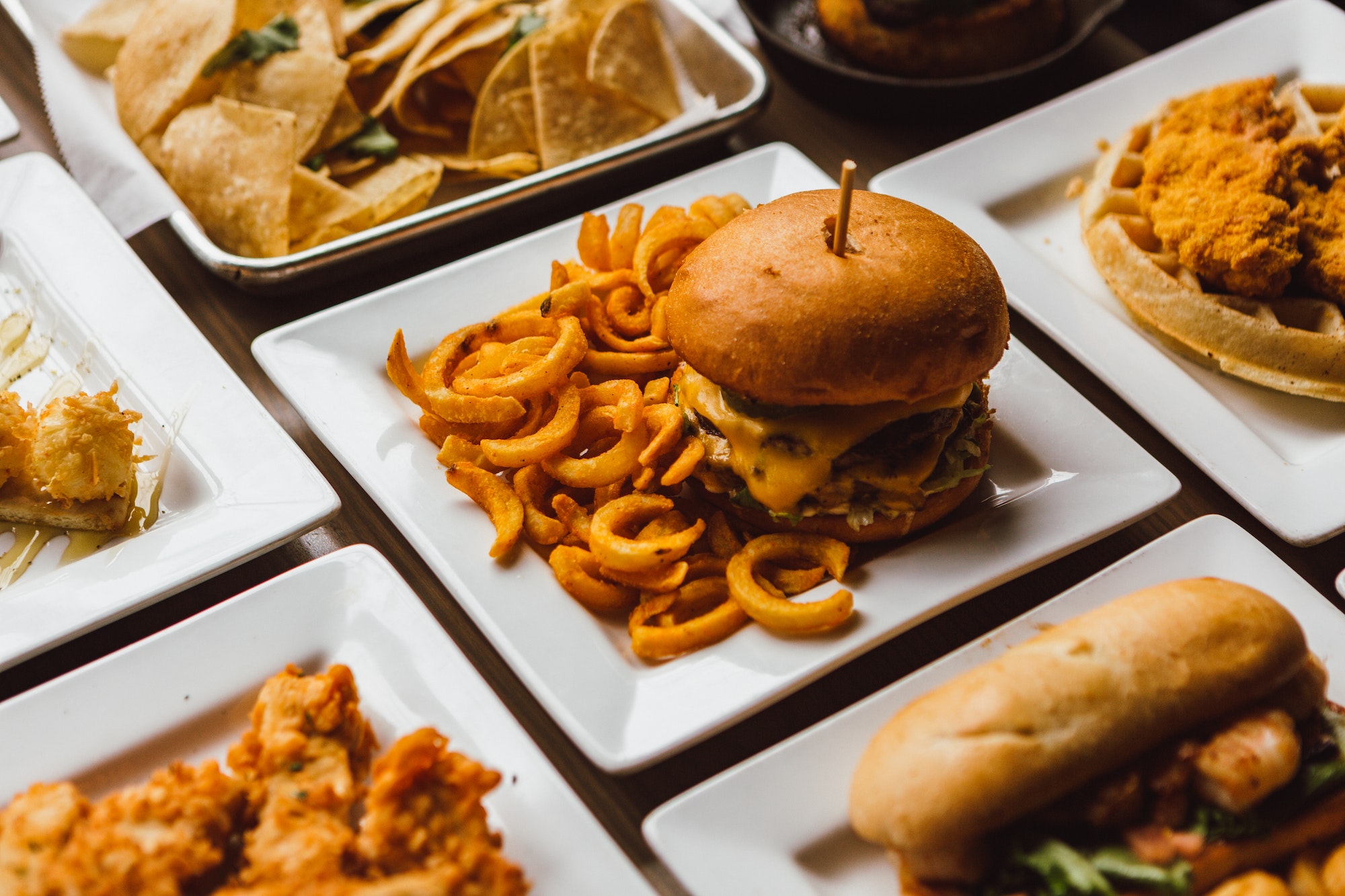 Restaurant table with square white plates full of yummy food; burgers and fries, chicken and waffles